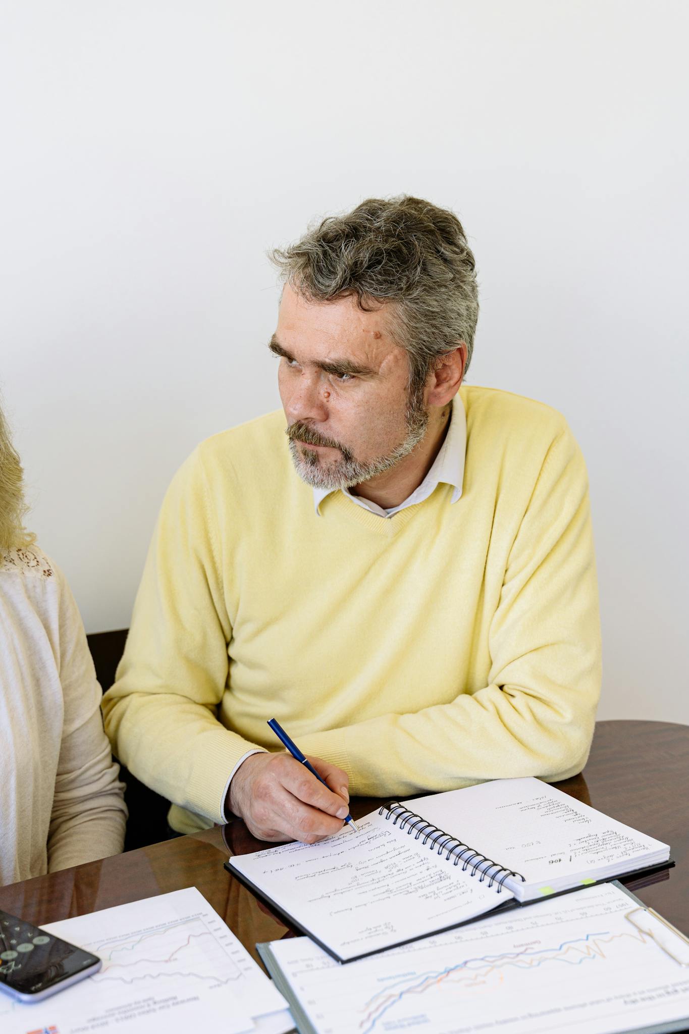 Elderly man in a yellow sweater writing with a pen in a notebook, seated at a desk.